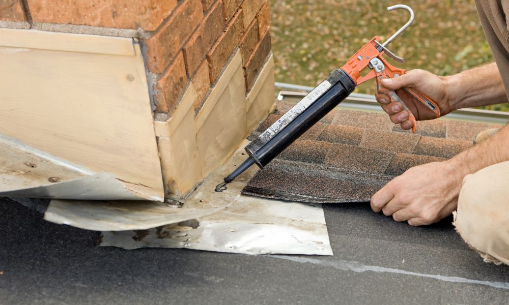 A close-up of a contractor using a caulking gun to apply sealant around the metal roof flashing at the base of a brick chimney during a roof repair.