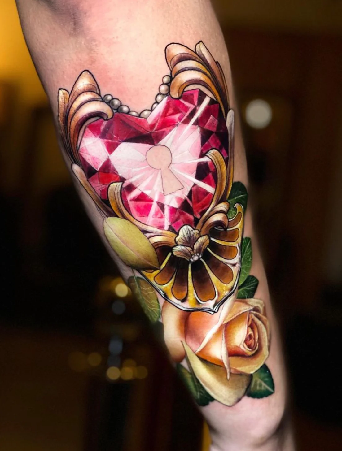 A vibrant, Neo-Traditional style tattoo on an arm featuring a deeply saturated, faceted red gemstone shaped like a heart with a keyhole at its center. The jewel is framed by ornate gold filigree scrollwork, a pearl border, and an elegant yellow rose with green leaves at the base.