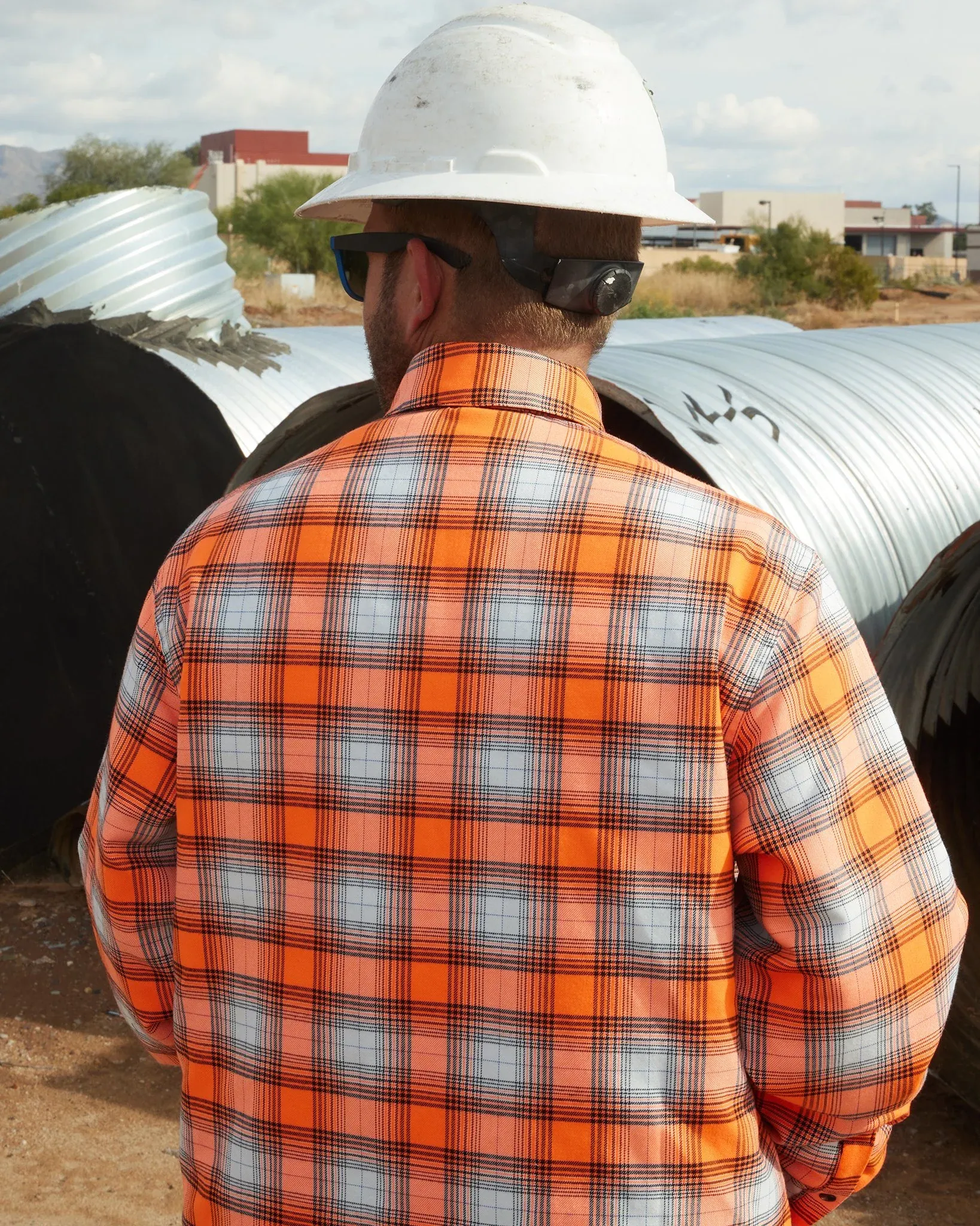 Back view of a worker in a hard hat wearing a high-visibility orange and gray plaid flannel jacket on a job site.