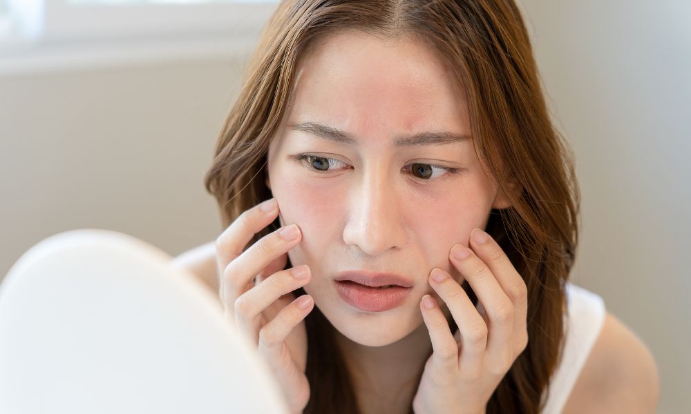 A woman looking concerned in the mirror at her skin, which shows signs of redness, highlighting the need to use the best tallow sunscreen for sensitive skin.