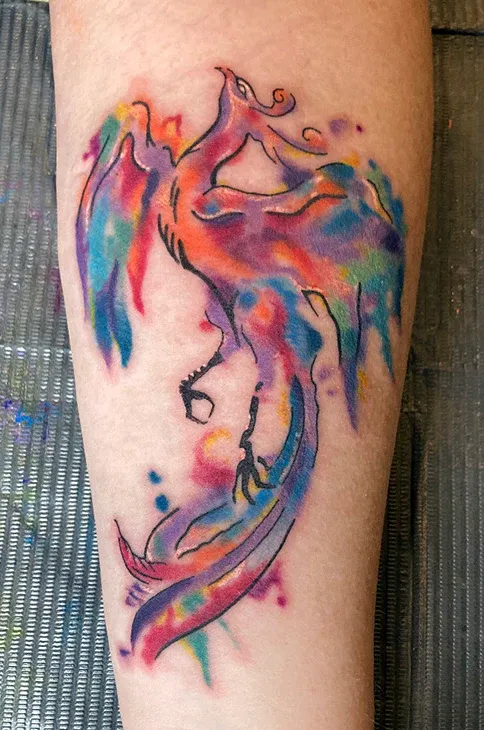 Vibrant watercolor tattoo of a phoenix with colorful splashes of blue, purple, red, and orange.