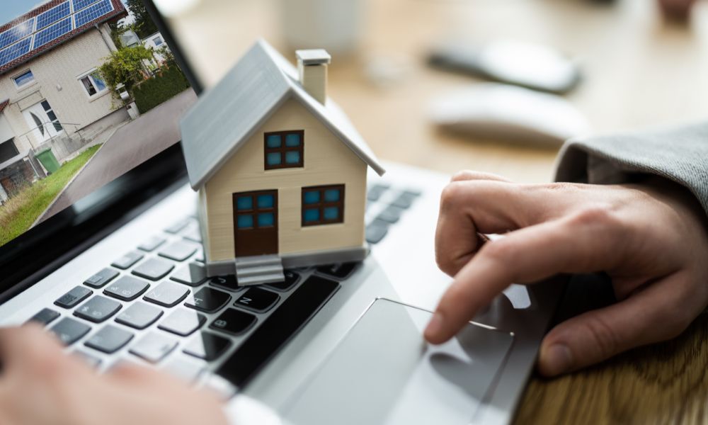 A small model house sitting on a laptop keyboard as a person uses the trackpad to view a luxury home listing on the screen.