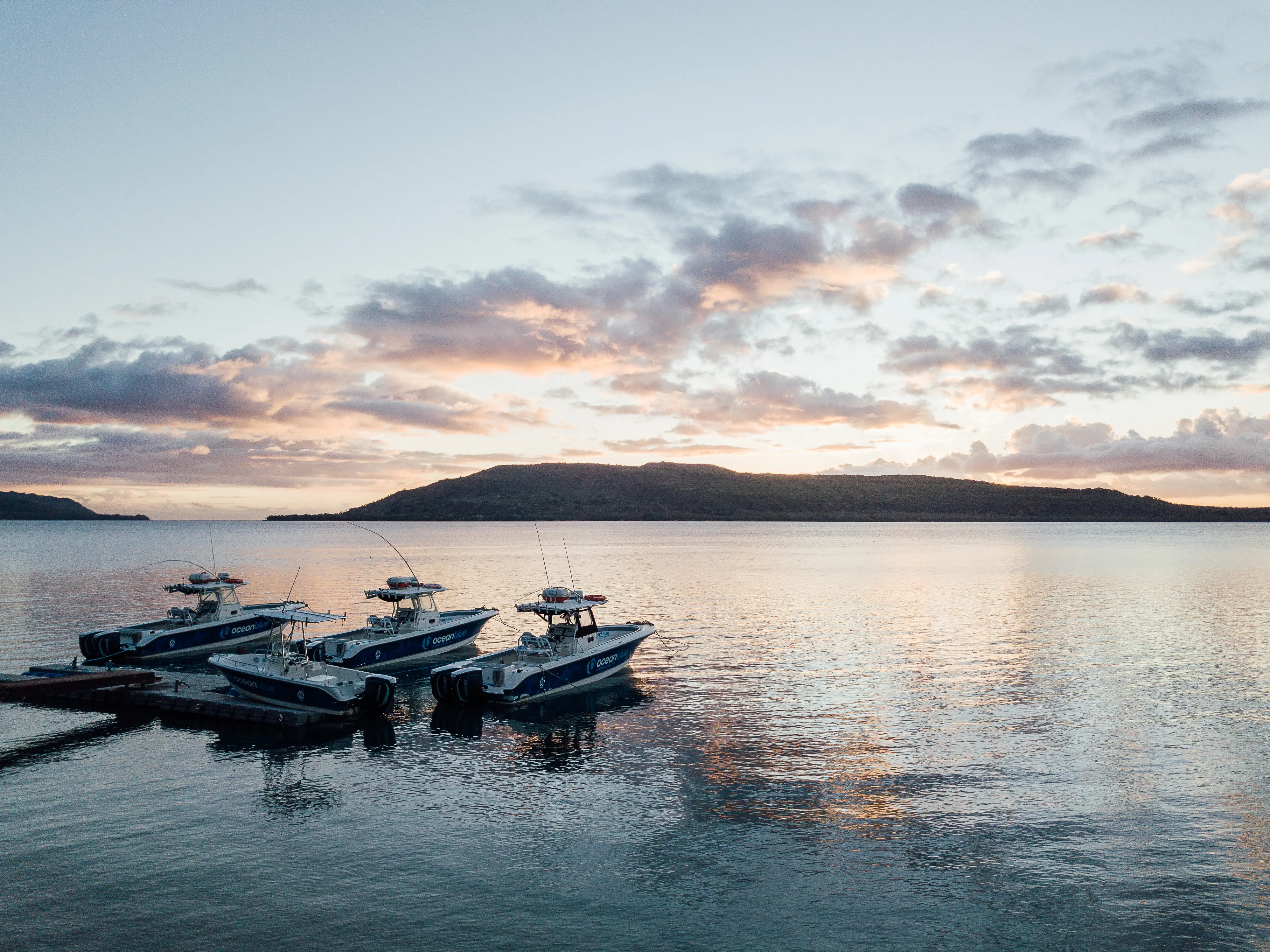 Ocean Blue Fishing Adventures boats moored at a jetty on calm water at sunset in Vanuatu, with island silhouettes and pastel clouds in the sky.