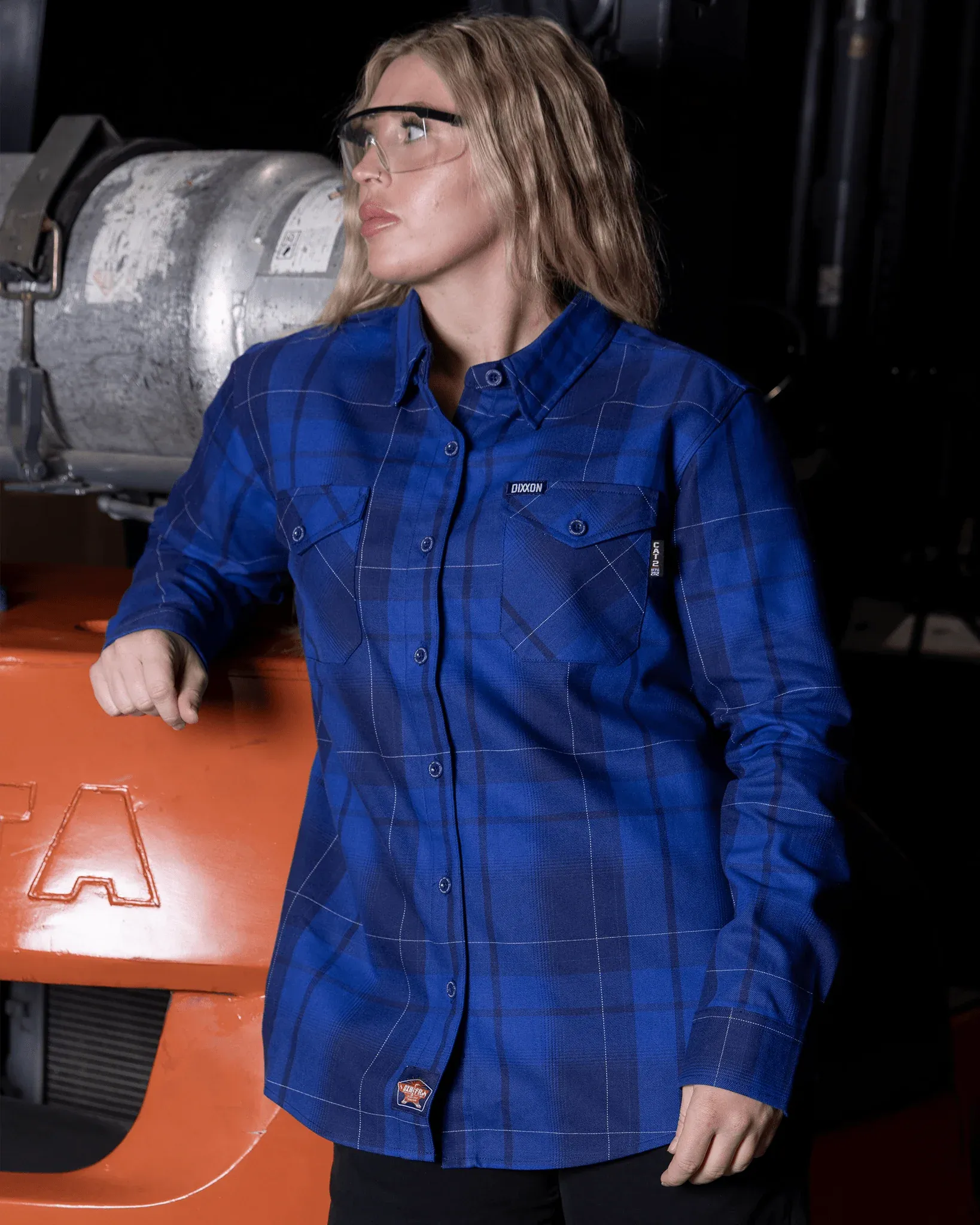 Woman wearing the royal blue Punch List FR Dixxon flannel shirt posing with safety glasses in an industrial setting.