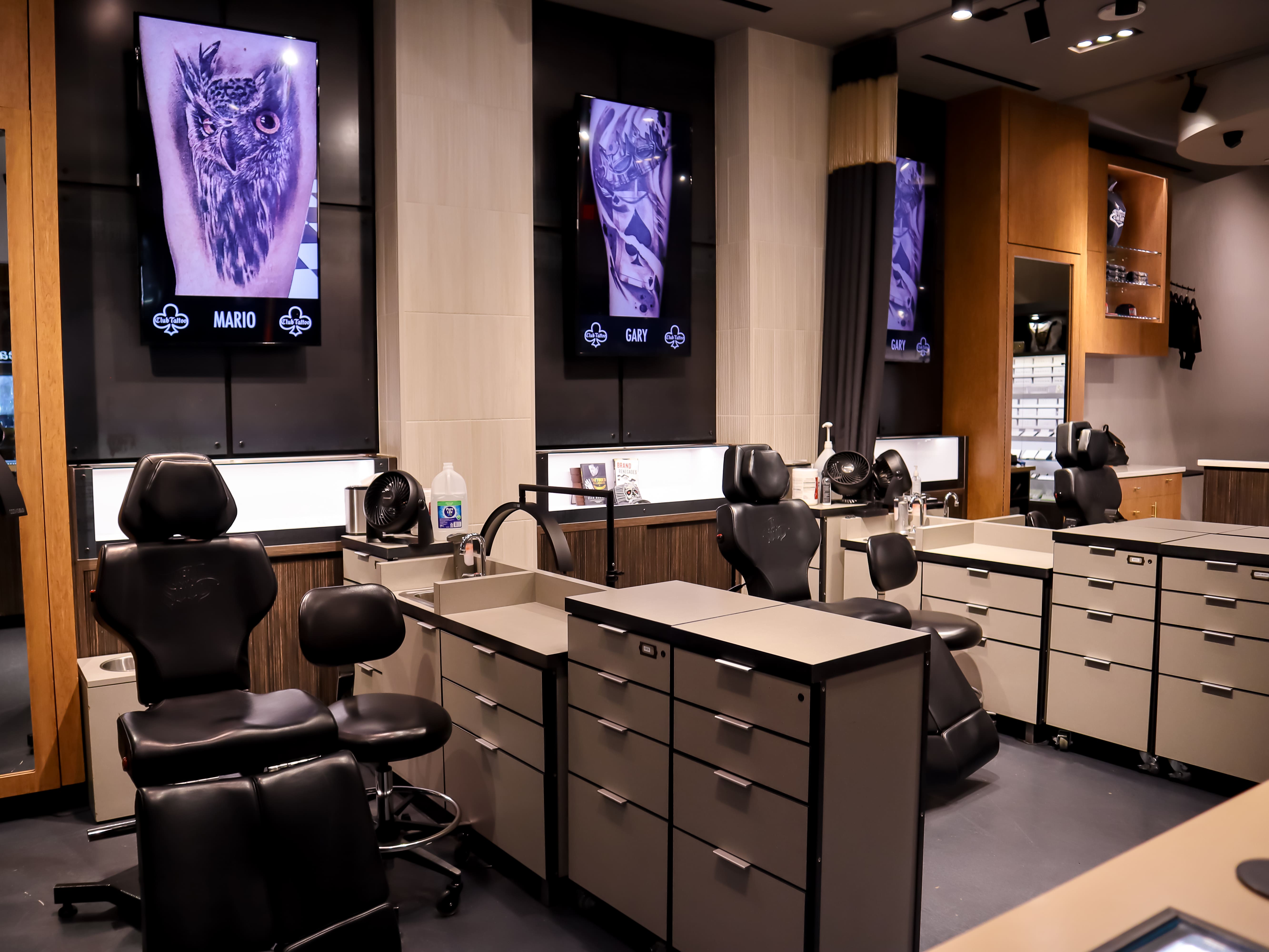 A clean, modern luxury tattoo studio interior featuring black leather adjustable client chairs, rolling stools, sleek grey counter stations with drawers, individual hand sinks, and digital screens displaying artist names and portfolio work.