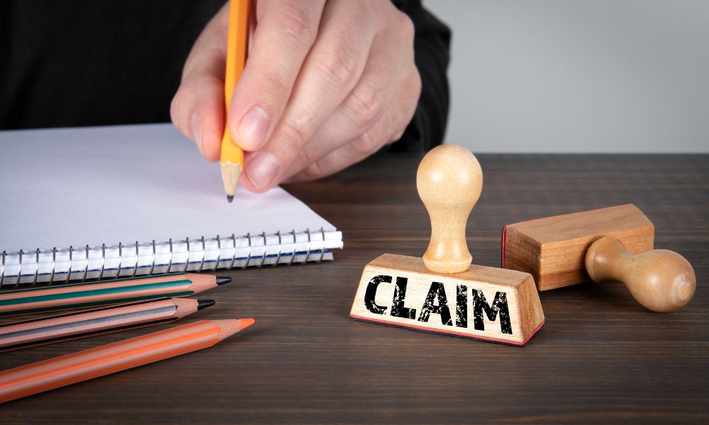 A close-up of a person writing documentation next to a wooden stamp that reads CLAIM, illustrating the official process of filing a coverage request under new home warranties.