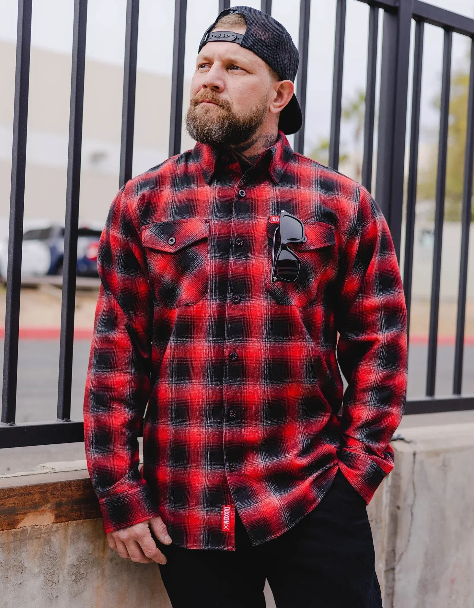 Bearded man in a backward cap wearing the red and black Fairview plaid Dixxon flannel with sunglasses tucked in pocket.