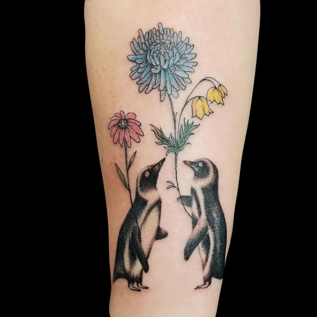 An illustrative tattoo on a forearm featuring two small, realistic black and gray penguins looking up at a tall, slender stem with a large light-blue chrysanthemum flower, a stalk of yellow bell-shaped flowers, and a single pink daisy