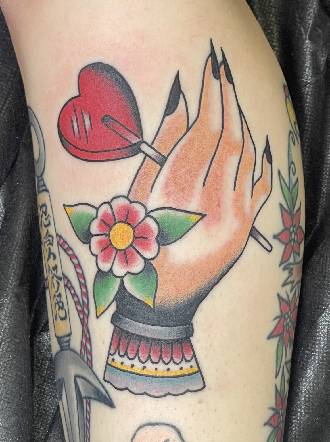 A person's inner bicep or thigh features a bold traditional hand tattoo. The hand has black nail polish, and a simple five-petaled flower sits on its wrist. The hand is holding up a stylized red, heart-shaped lollipop.