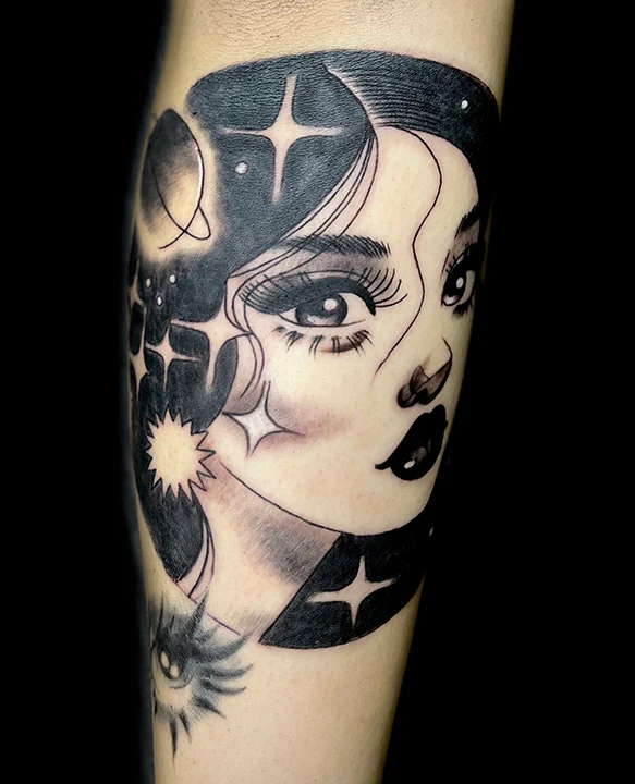 A black and grey illustrative tattoo of a woman's face. Her hair is filled with a solid black space-themed pattern featuring a planet, multiple stars, and a sun, with a stylized eye design visible at the bottom.
