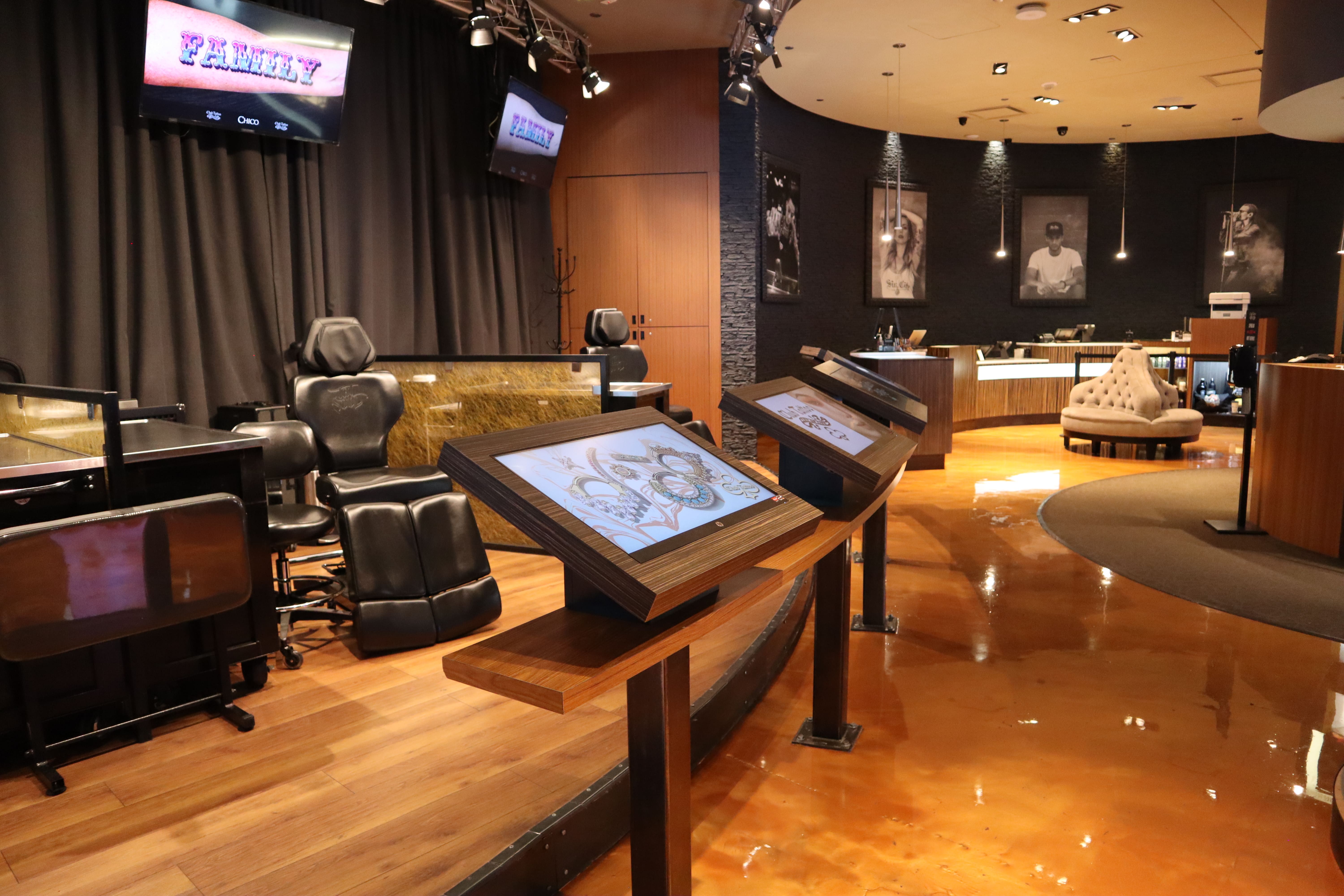 The interior of a modern Club Tattoo studio displaying several patented Interactive Tattoo Touchscreens on wooden stands for client browsing, situated next to adjustable black leather tattoo chairs, workstation counters, and overhead screens reading "FAMILY" against dark curtains.