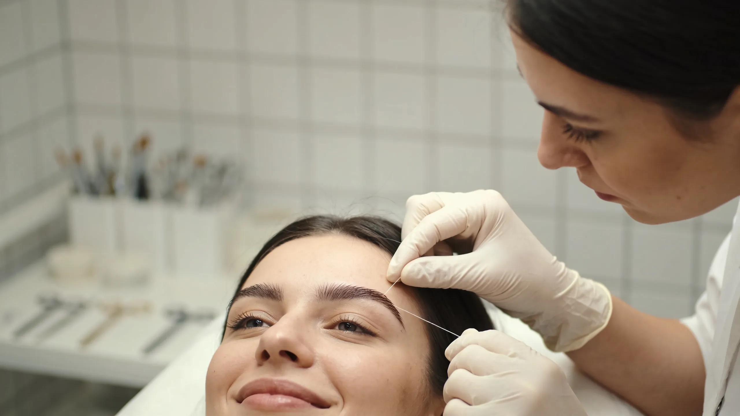 Aesthetician shaping eyebrows using threading techniques during a beauty treatment