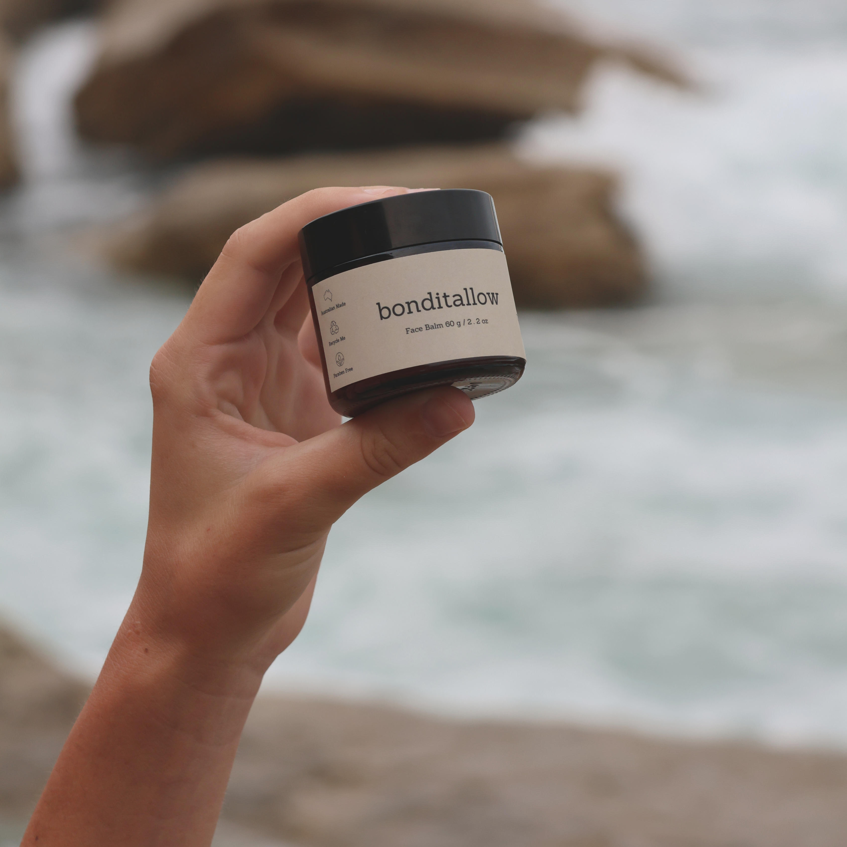 Hand holding a jar of Bonditallow skincare product near the ocean