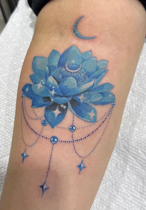 Luminous blue lotus flower tattoo adorned with hanging bead chains and crescent moons.