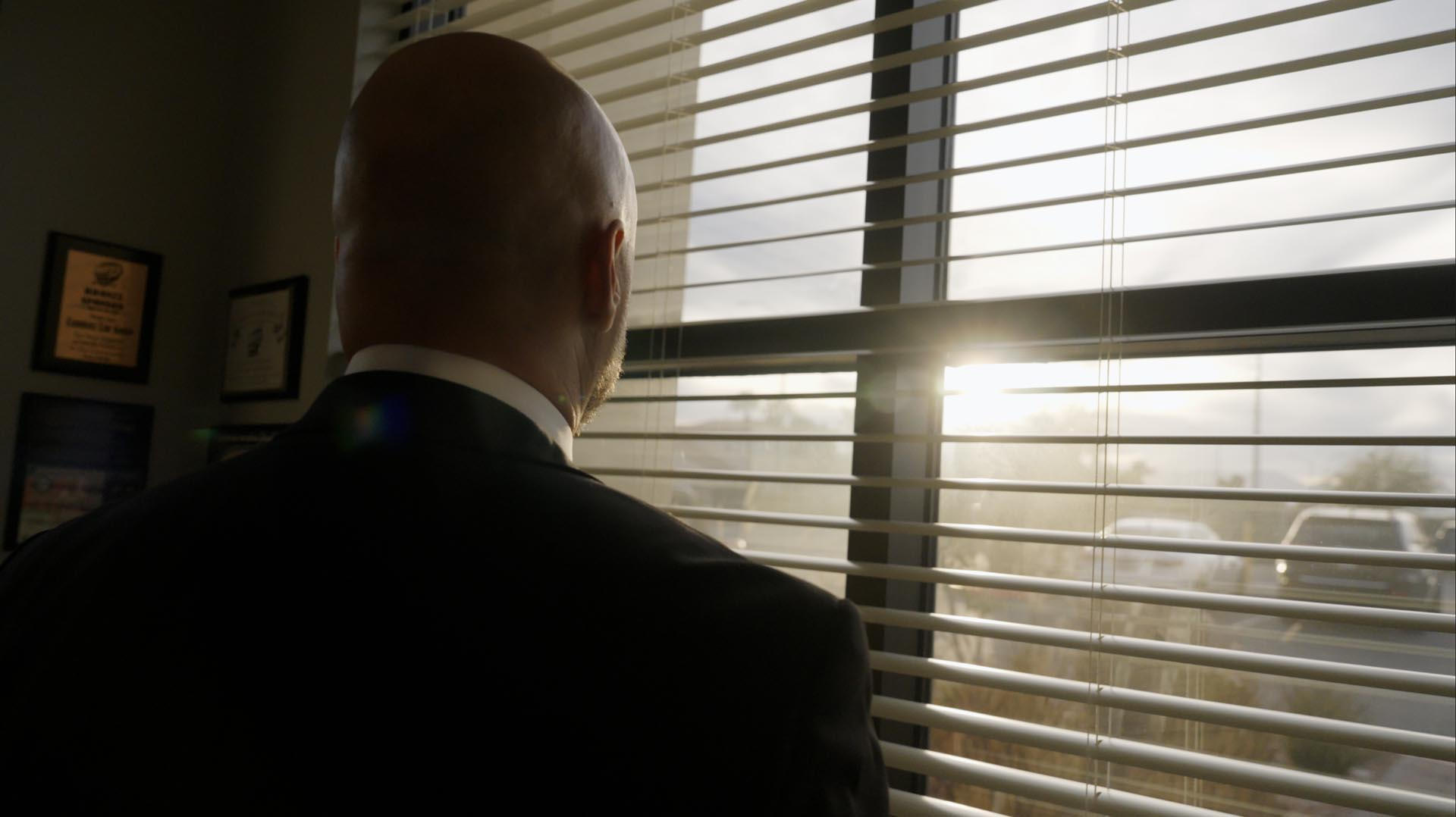 Man in a suit looking out a window with blinds, sunlight shining through