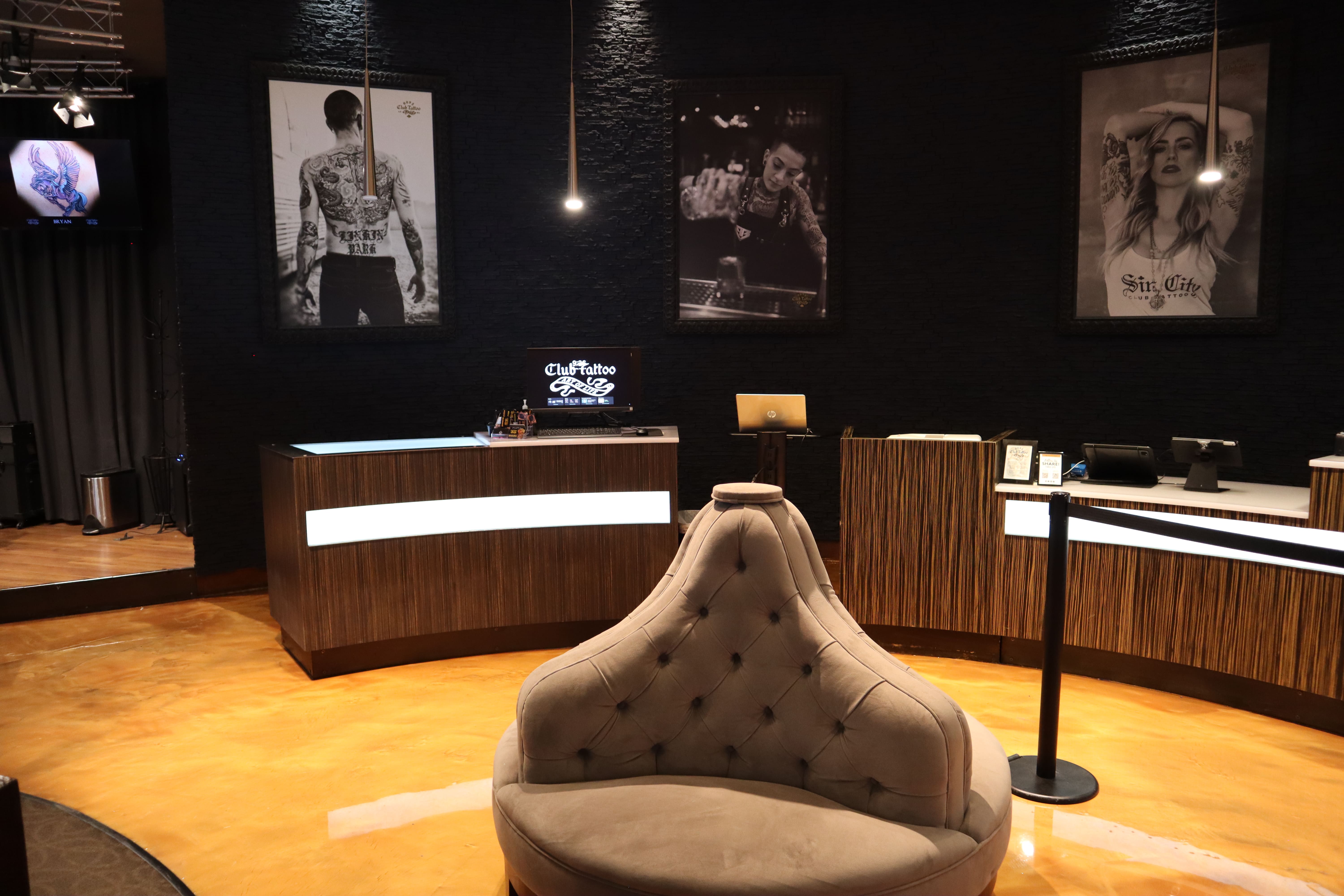 A luxury lounge and reception area at Club Tattoo, featuring a plush tufted circular seating chair, dark textured brick walls adorned with large black-and-white portraits of tattooed individuals, and modern wood-paneled service counters with integrated light panels under warm ambient lighting.