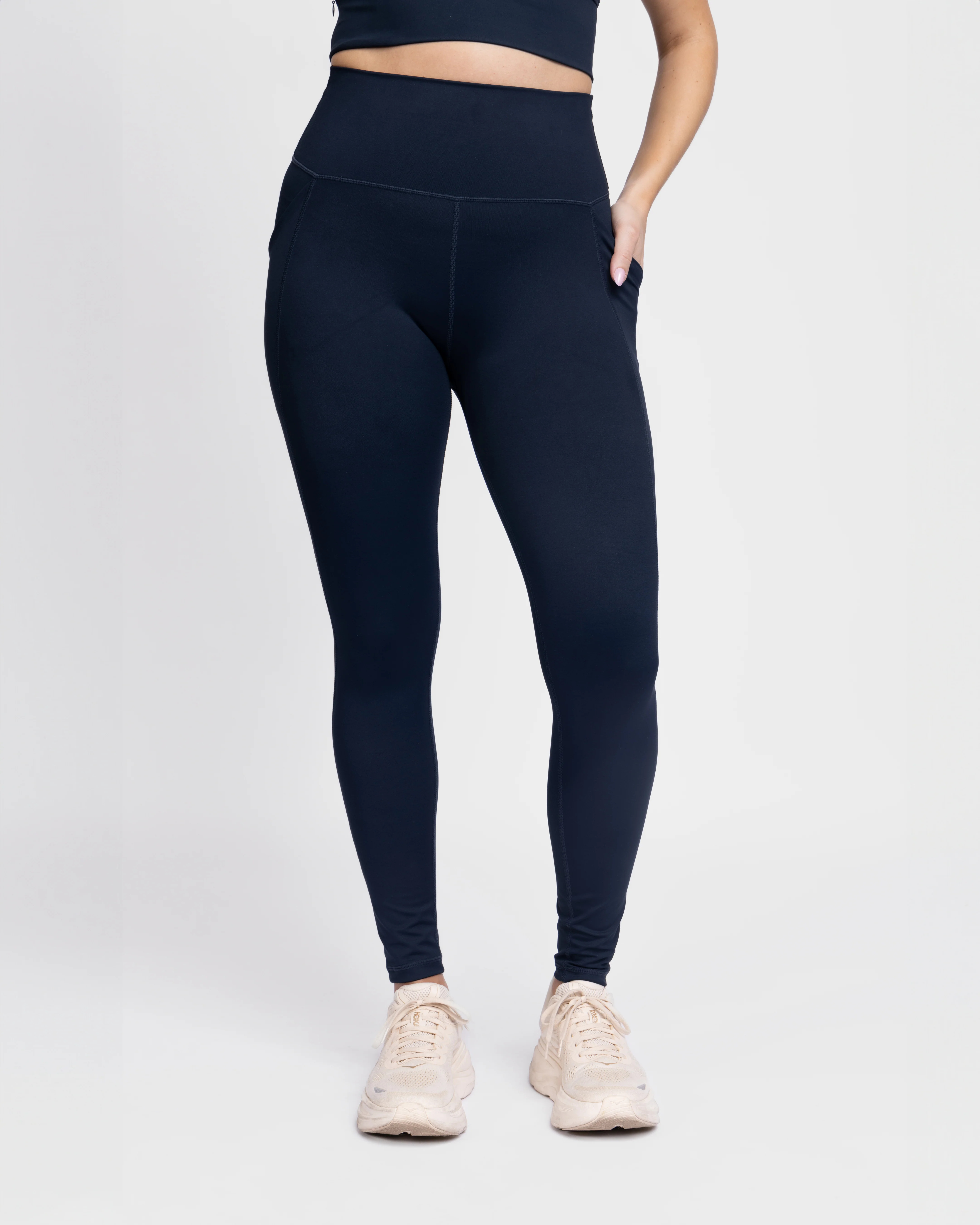 Lower-body view of a model standing in high-waisted navy blue workout leggings featuring a side pocket on the hip, paired with light cream running sneakers.