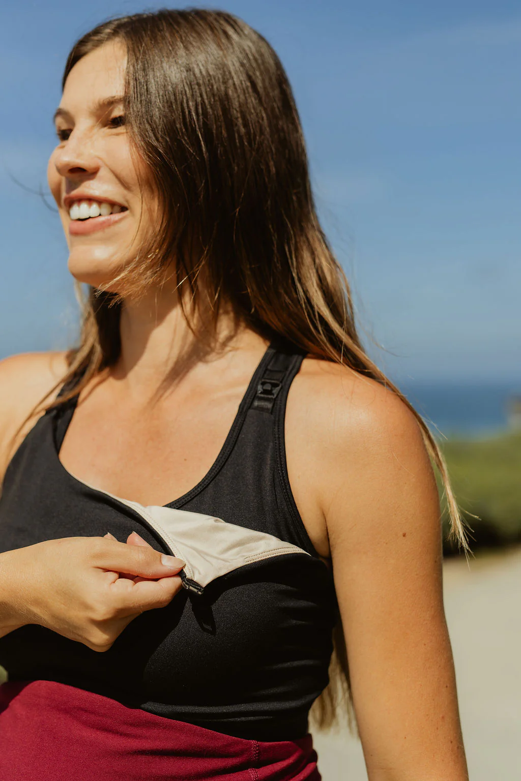 Outdoor lifestyle photo of a smiling woman wearing a black Senita maternity tank top and maroon bottoms, showing the easy-access drop-down nursing cup feature with a tan inner lining against a soft-focus blue sky background.