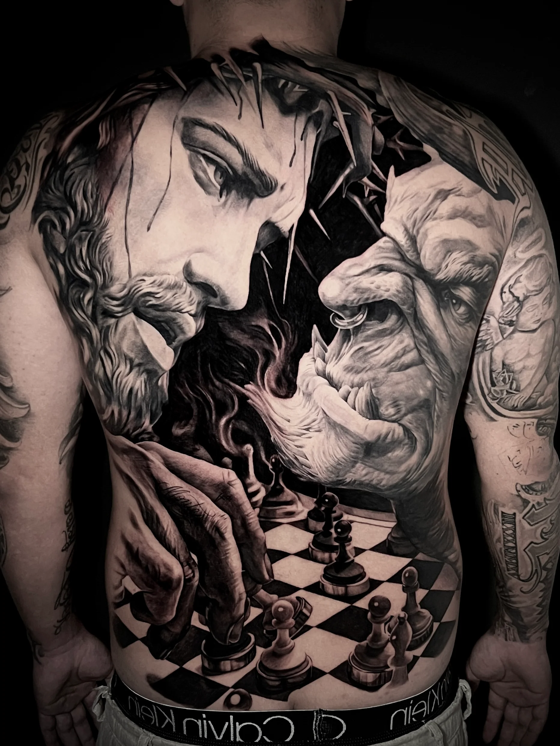 A massive, full-back black-and-grey realism tattoo illustrating an intense chess match. On the left side, a detailed, bearded figure wearing a crown of thorns contemplates a move, while on the right side, a wrinkled, horned creature or troll watches closely from across the checkered board.