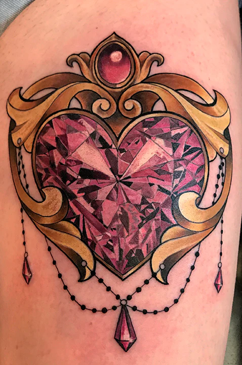 An ornate, illustrative tattoo of a large pink heart-shaped gemstone encased in a decorative gold frame with hanging beads and smaller teardrop jewels.