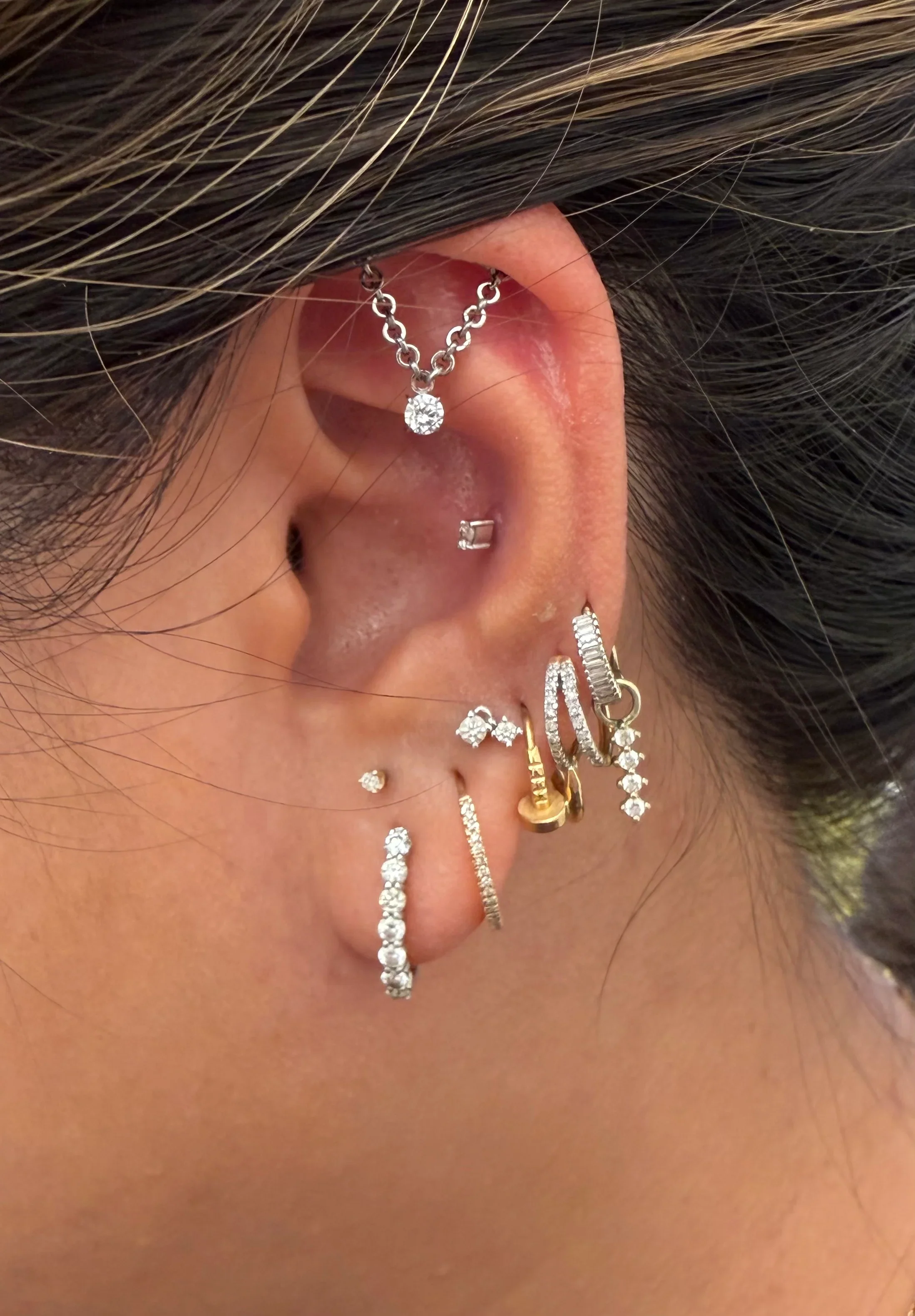 Detailed side profile of a complex, curated ear constellation project featuring multiple silver and gold layered hoops in the earlobe, a flat helix stud attached to a hanging silver chain accent, and a simple stone stud in the lower conch.