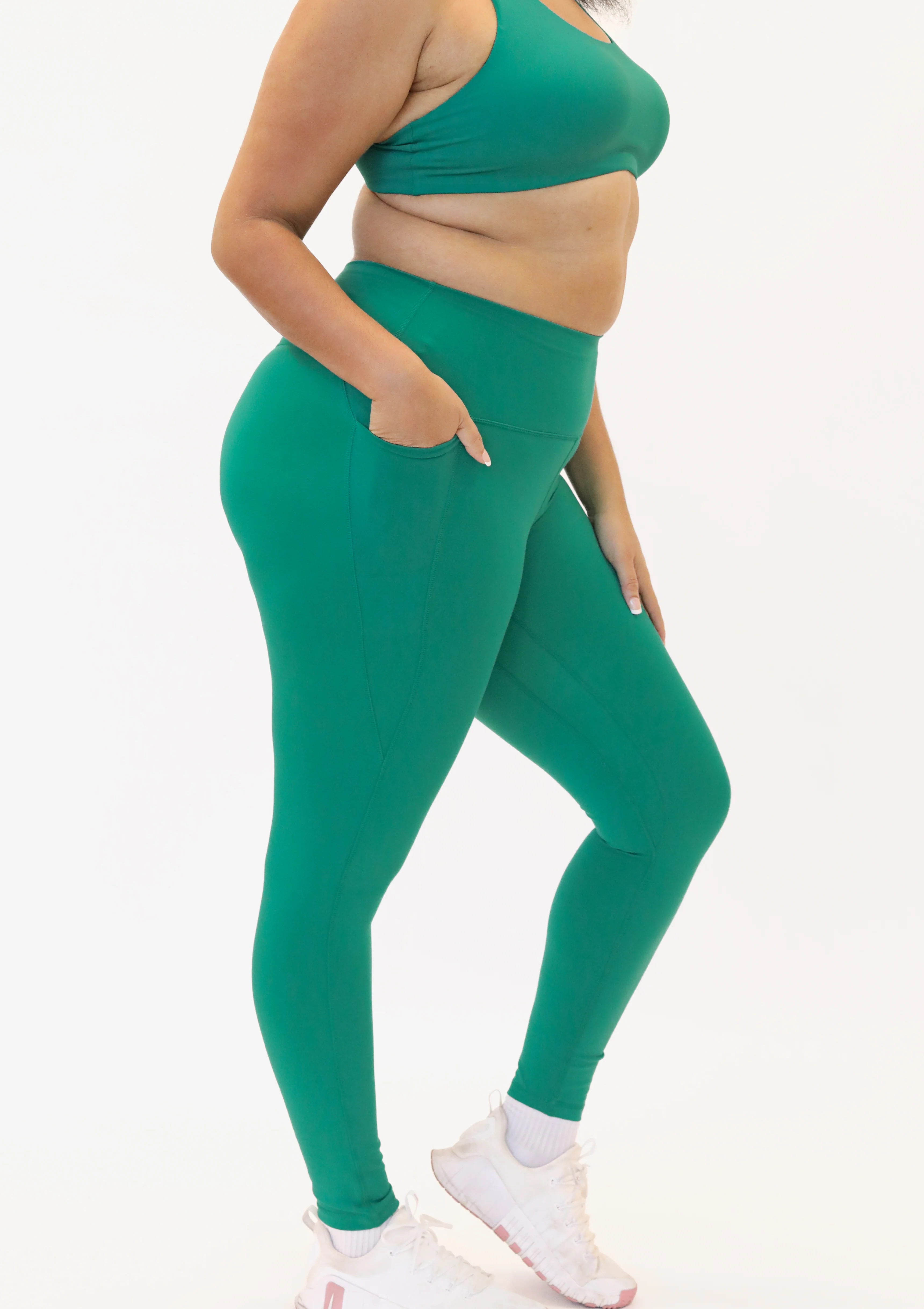 Side view of a woman wearing high-waisted activewear leggings and a matching sports bra in a vibrant viridian green color. She has her hand resting inside a deep side pocket built into the thigh of the leggings.