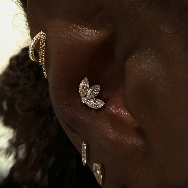 Close-up of a curated ear piercing design on dark skin featuring a gold conch stud shaped like a three-leaf cluster with inlaid marquise-cut diamonds, paired with a diamond-studded forward helix hoop with a delicate hanging gold chain.