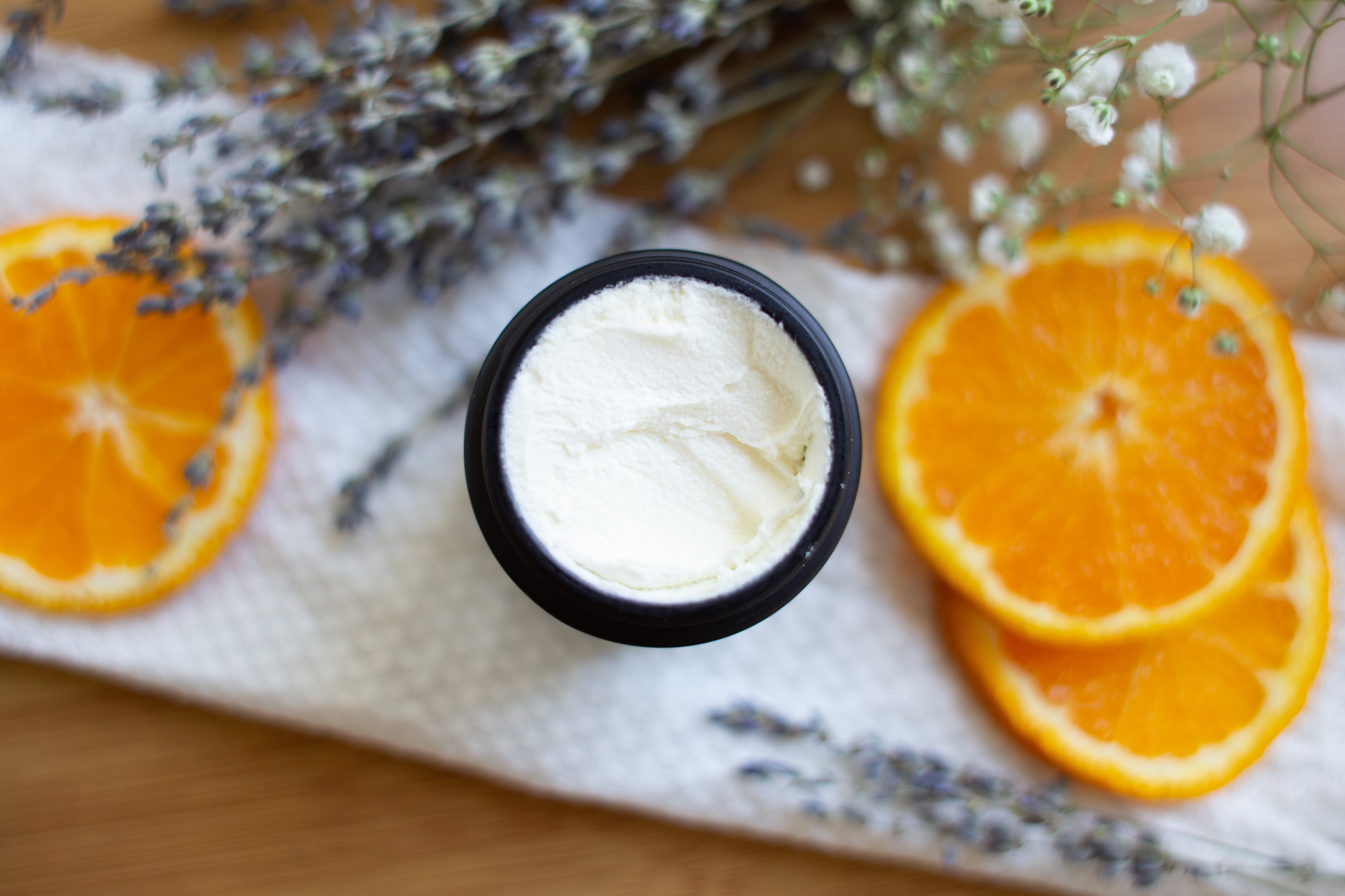 An open jar of whipped white beef tallow for hair surrounded by fresh orange slices, dried lavender, and baby's breath, highlighting natural ingredients in hair growth products.