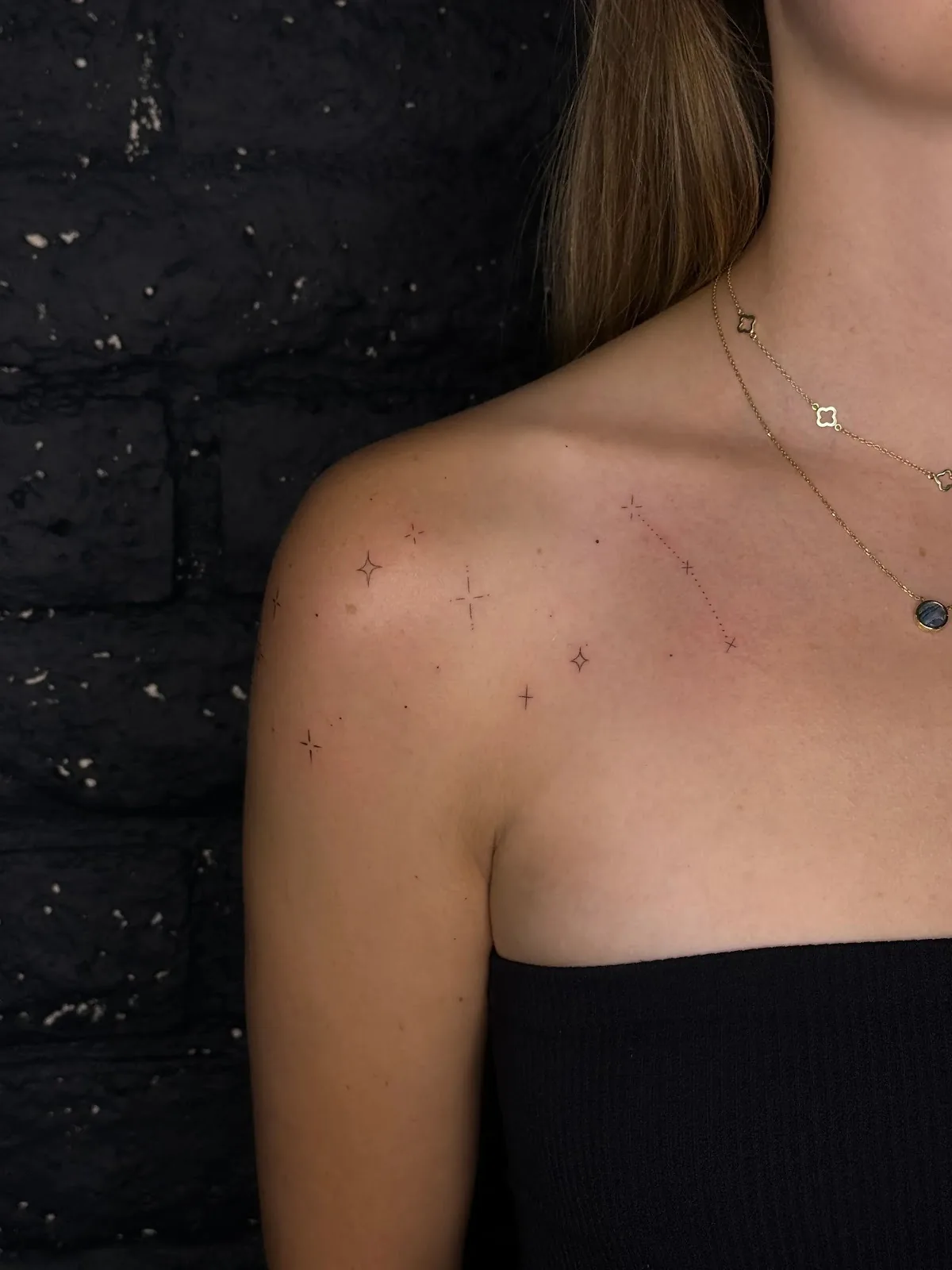 Delicately tattooed tiny stars and constellation sparkles scattered across a woman's shoulder and collarbone.