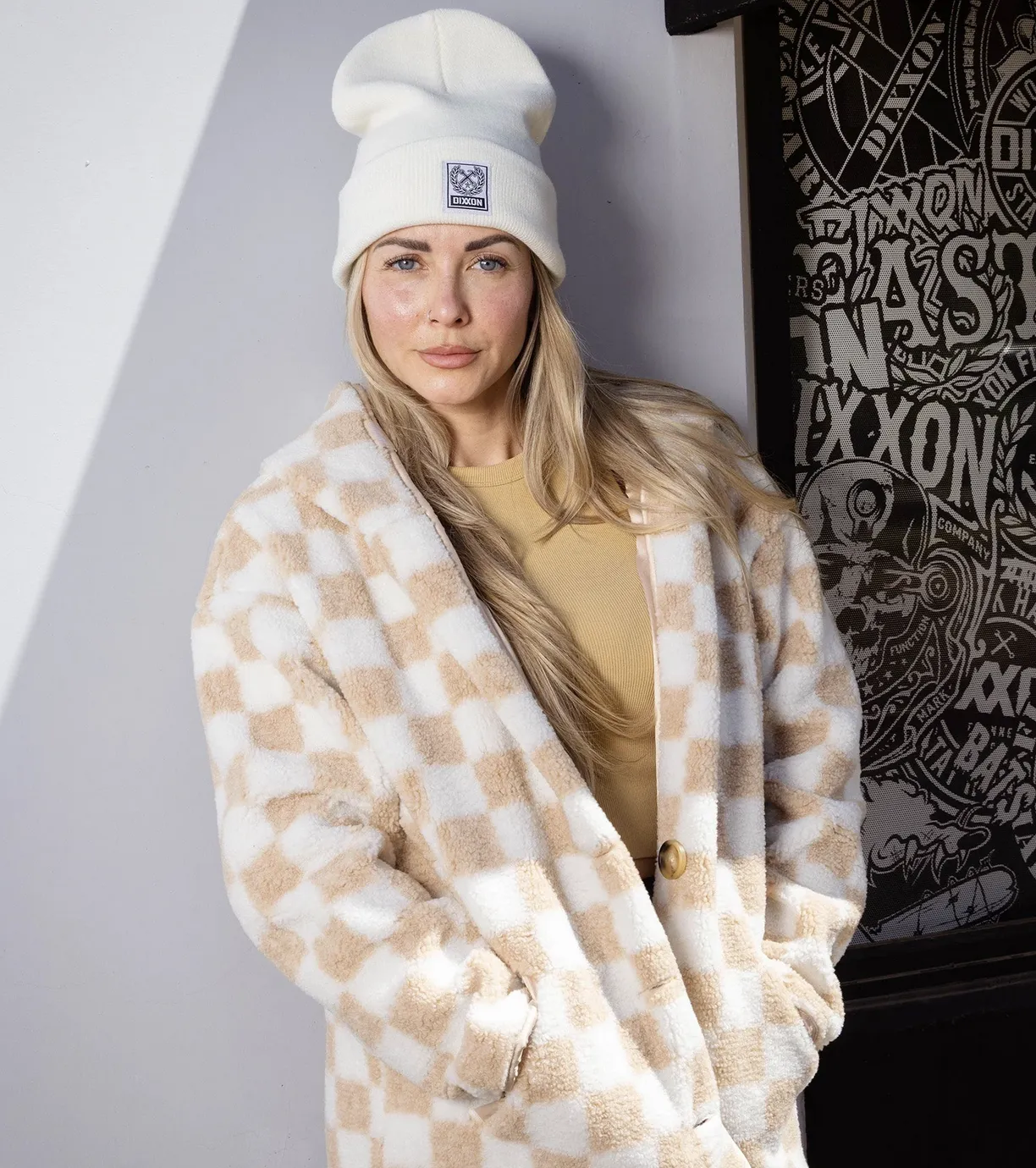 A woman with blonde hair and a white beanie stands against a white wall wearing a long, plush cream and tan checkered sherpa coat. She is looking directly at the camera with her hands in the coat pockets.