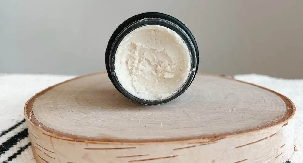 Overhead close-up of a small, open black jar filled with a light, creamy balm, resting on a round birch wood slice, symbolizing natural elements in a dedicated skincare routine.