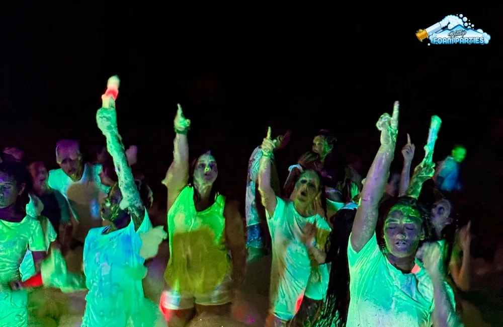 A group of excited people at a foam glow party in nashville, dancing and raising their hands in the dark, illuminated by neon glow light and covered in brightly colored foam.
