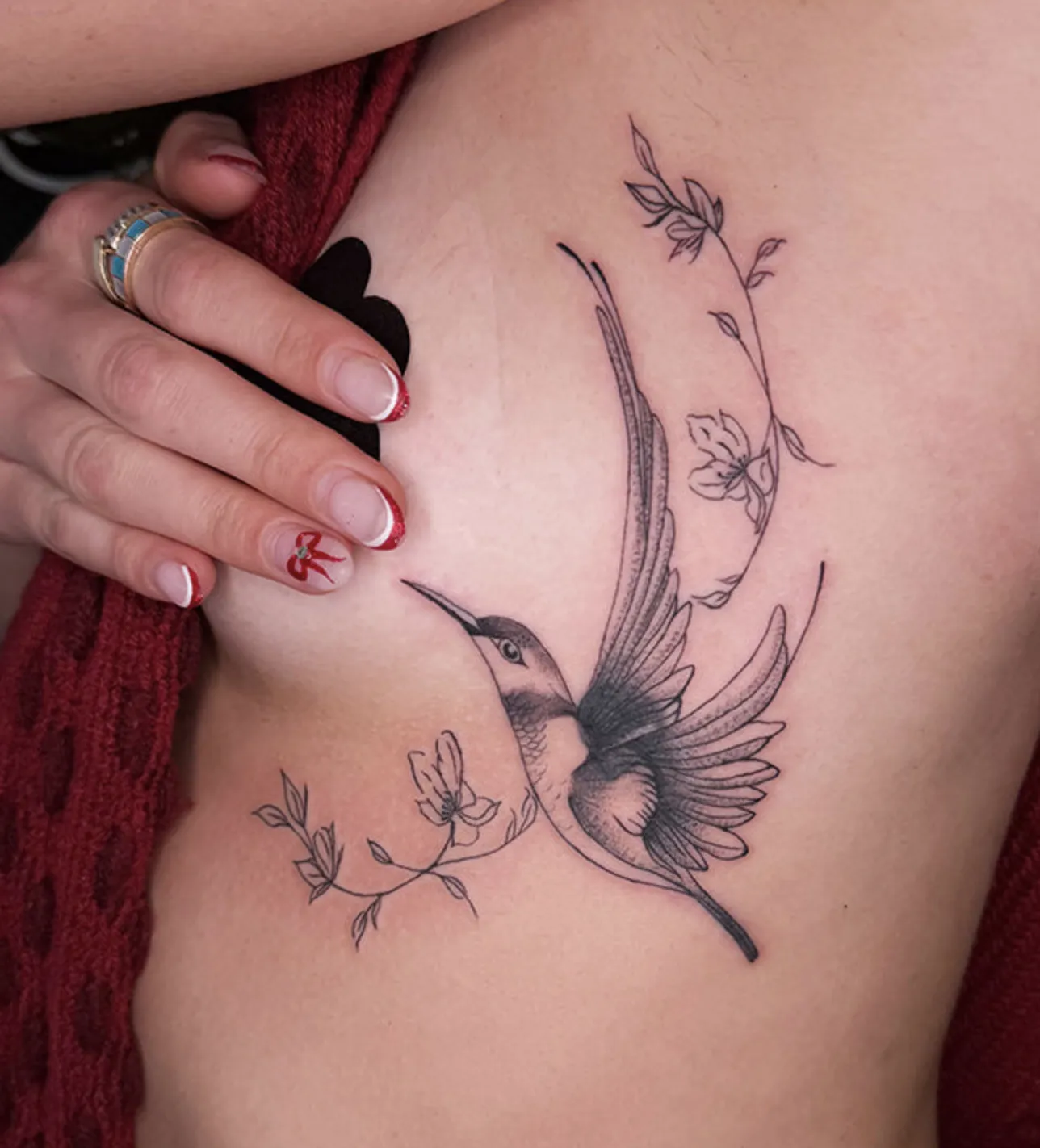 Delicate fine-line tattoo on the ribs of a hummingbird in flight, surrounded by thin, graceful floral branches and light stippled shading.