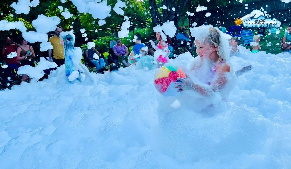 A young girl smiles while holding a colorful beach ball and playing in a thick layer of bubbles during a foam event. Other children are visible in the background, also covered in bubbles, while adults watch from the side during one of many successful foam parties. The hilltop foam parties logo is displayed in the upper right corner.