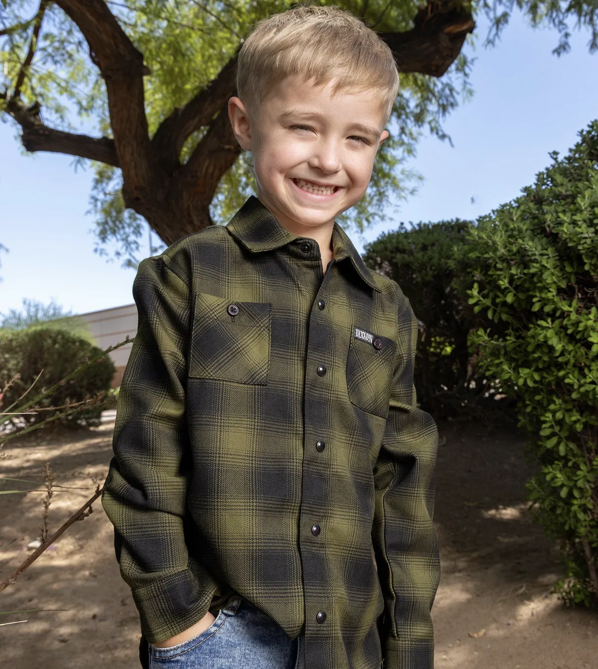 Young boy in green outdoor setting smiling while wearing Dixxon Lone Wolf Flannel