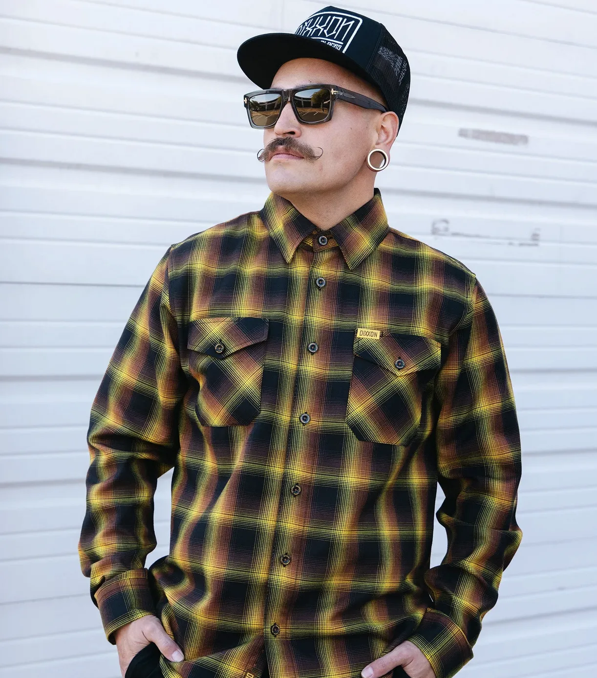 A man with a mustache and gauge earrings wearing a yellow, black, and orange plaid Dixxon D-Tech™ flannel shirt with a small yellow logo tag on the chest pocket. He is also wearing a black trucker hat with a white Dixxon text logo and dark sunglasses.