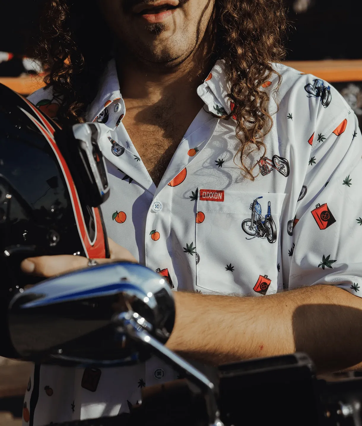 A close-up of a person wearing the white Dixxon "Free Ride" party shirt, which features a playful print of motorcycles, oranges, gas cans, and leaves. The person is holding a black motorcycle helmet, highlighting the brand’s connection to biker culture