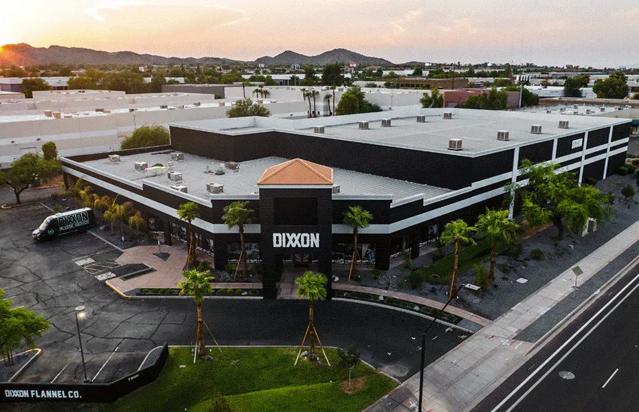 An aerial sunset view of the expansive Dixxon Flannel Co. headquarters and showroom. The modern building features bold black exterior walls with stark white architectural lines, surrounded by palm trees and a branded Dixxon mobile unit in the parking lot.