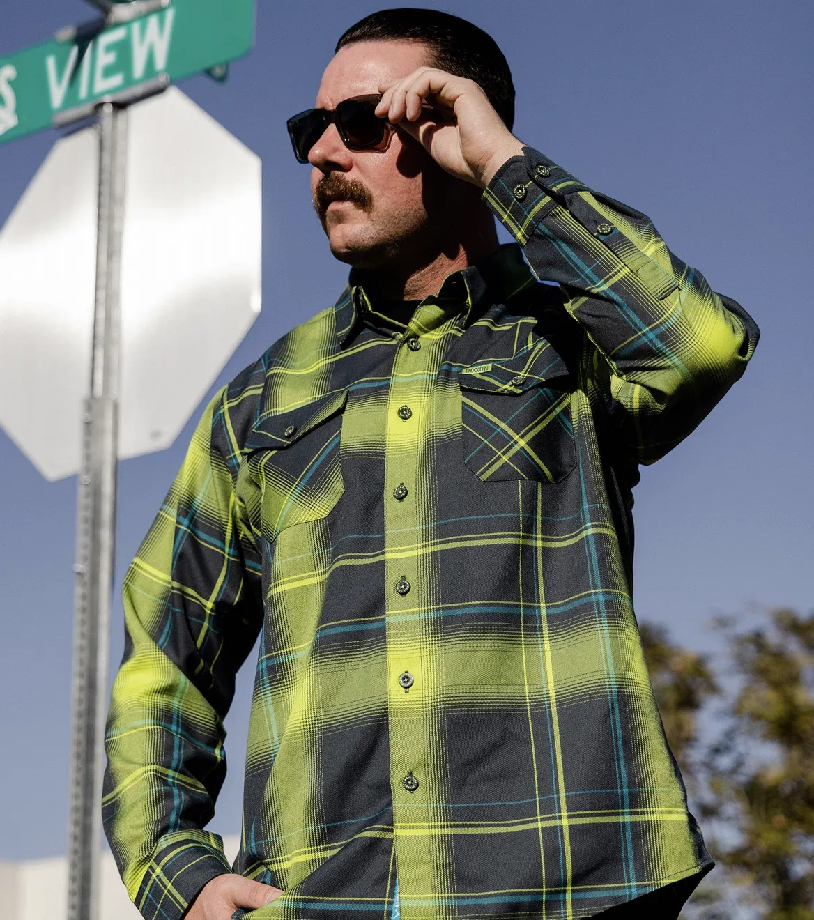 A close-up of a man in sunglasses and a mustache wearing a vibrant lime green and charcoal gray plaid flannel, featuring the signature Dixxon D-Tech™ blend and mitered edge cuffs.
