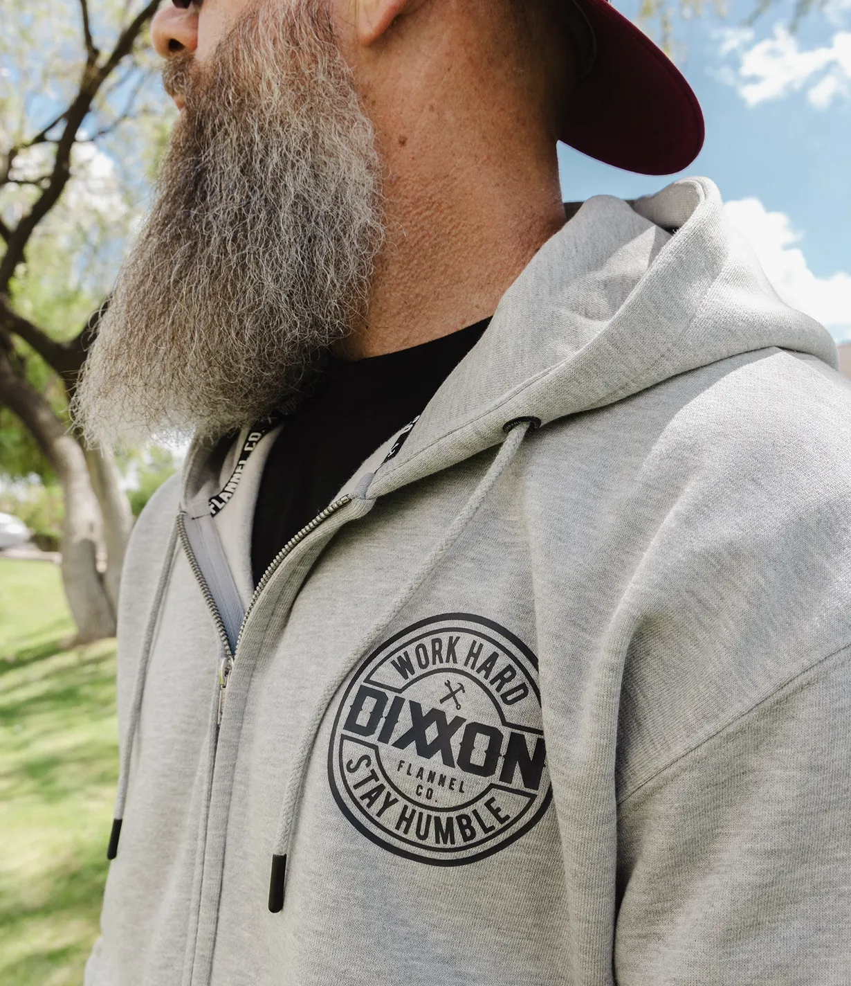 Close-up of a grey zip-up hoodie featuring a circular Dixxon Flannel Co. "Work Hard Stay Humble" logo on the chest and branded taping inside the neck.