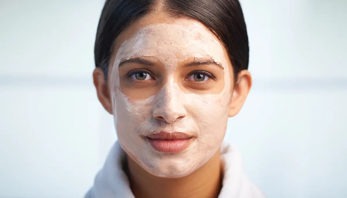 A young woman wearing a white robe with a thick, cream-colored facial mask applied to her face as an oily skin treatment. The mask, which could be a product of tallow skincare, covers her forehead, cheeks, and chin, leaving the area around her eyes and mouth clear.