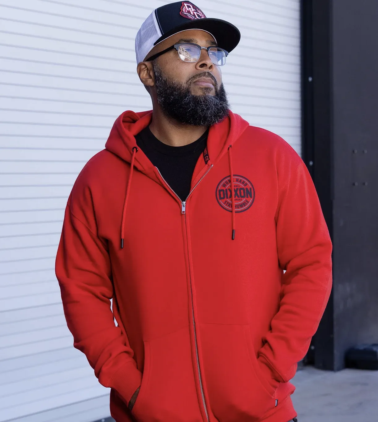 A man with a grey beard and glasses wearing a bright red zip-up Dixxon hoodie with a circular "Work Hard Stay Humble" corpo logo patch on the chest. He is also wearing a black and white mesh-back trucker hat featuring a red shield logo.