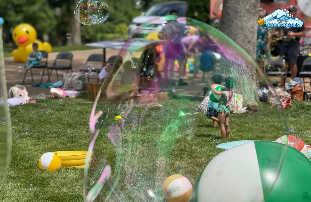 A large, iridescent bubble framing a lively outdoor park scene with children, beach balls, and an inflatable duck, capturing the excitement of bubble birthday parties in a sunny setting.