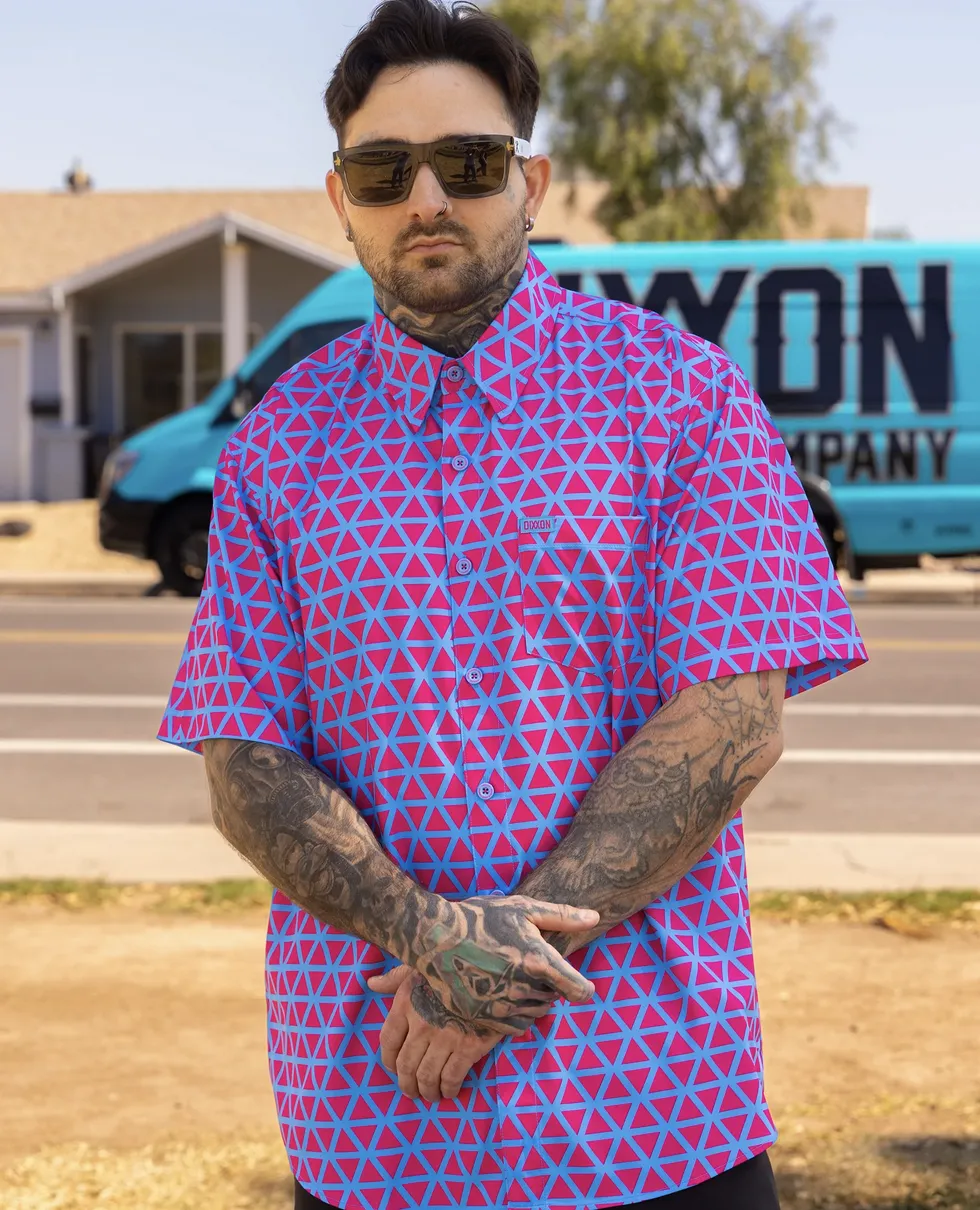 A man with neck and arm tattoos wears a vibrant short-sleeve button-down featuring a pink and cyan geometric triangle pattern. He stands in front of a blue Dixxon Company van with his hands folded.