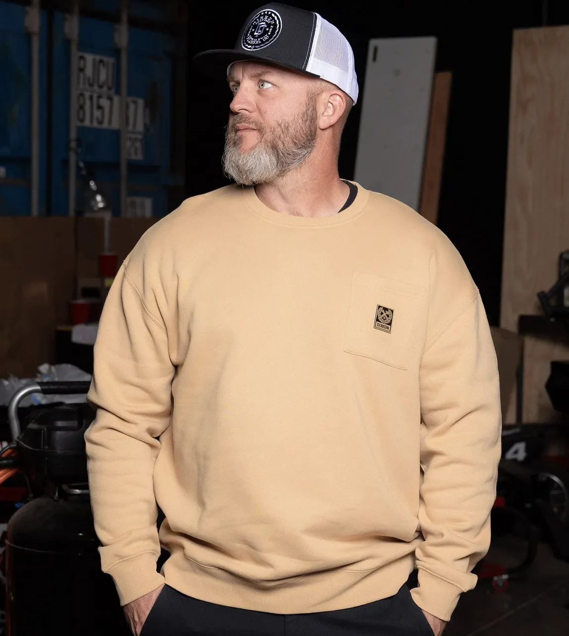 A man in a tan, fleece-lined Dixxon crewneck sweatshirt featuring a clean, rectangular logo patch on the front pocket. The relaxed fit and classic design are shown in a garage setting, representing the authentic, straightforward nature of the Dixxon community.