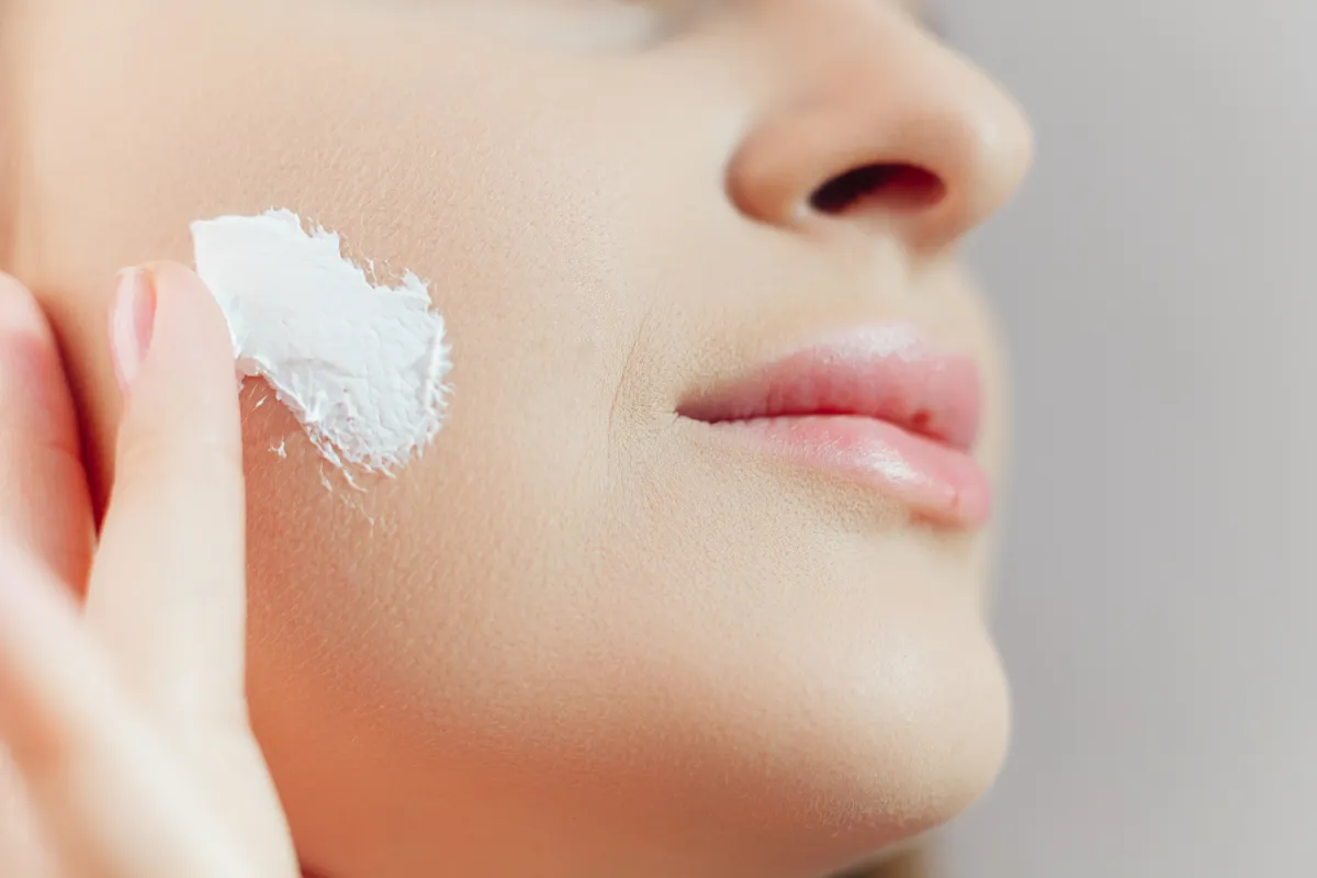 Close-up of a woman gently applying a thick white cream, such as Tallow Skincare balm, to her cheek.