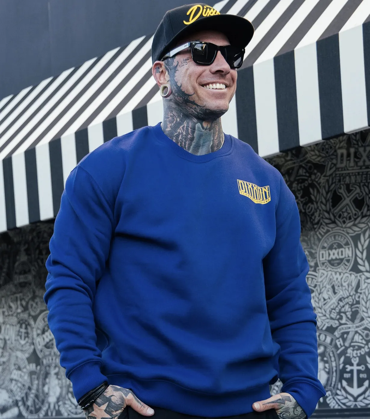 A man with neck and face tattoos wearing a vibrant royal blue crewneck sweatshirt with a gold Dixxon Flannel Company logo on the left chest.