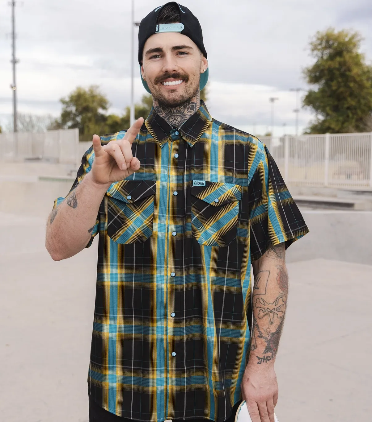 Man smiling, holding up the rock and roll hand sign, holding a skateboard, and wearing Dixxon Get Radical Short Sleeve