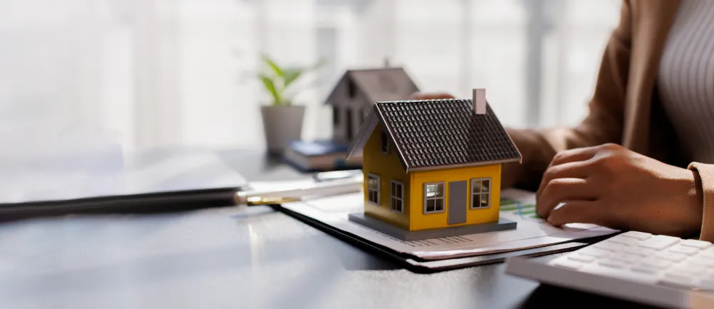 A luxury home agent finalizing a contract next to a yellow model house, symbolizing a successful closing for first-time home buyers during the home-buying process.