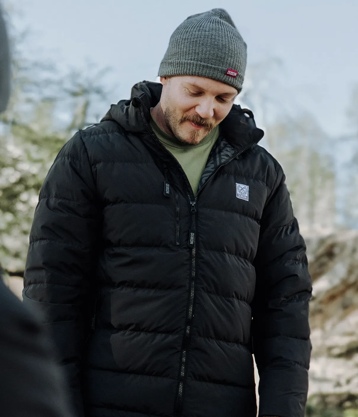 A person outdoors wearing a black Ridge Down Jacket with horizontal quilting and a branded logo patch on the left chest.
